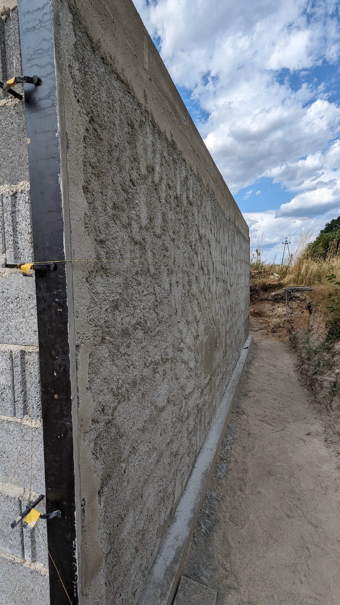 Ściana fundamentowa z bloczków betonowych, otynkowana szarą zaprawą, z widocznymi elementami poziomowania i wykopem w tle.