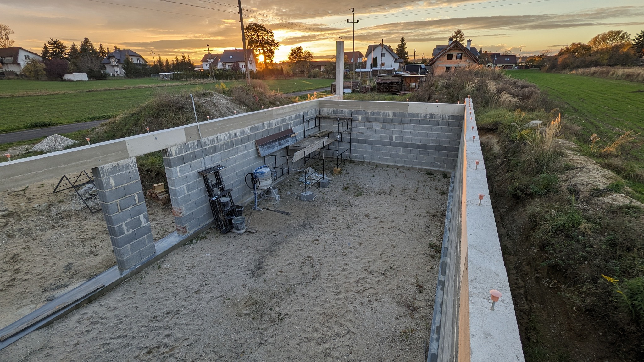 Fundamenty budynku z betonu komórkowego w trakcie budowy, widoczne szalunki i zbrojenia, na tle zachodzącego słońca nad polami.