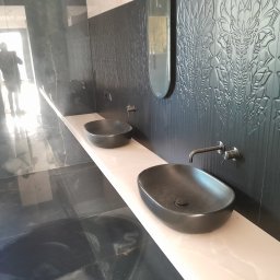 Andriy Derevyanko - Nowoczesna łazienka w Warszawie z dwoma czarnymi umywalkami na długim, jasnym blacie, ozdobna czarna ściana z motywem roślinnym i podłużnym lustrem, widoczna osoba w tle.