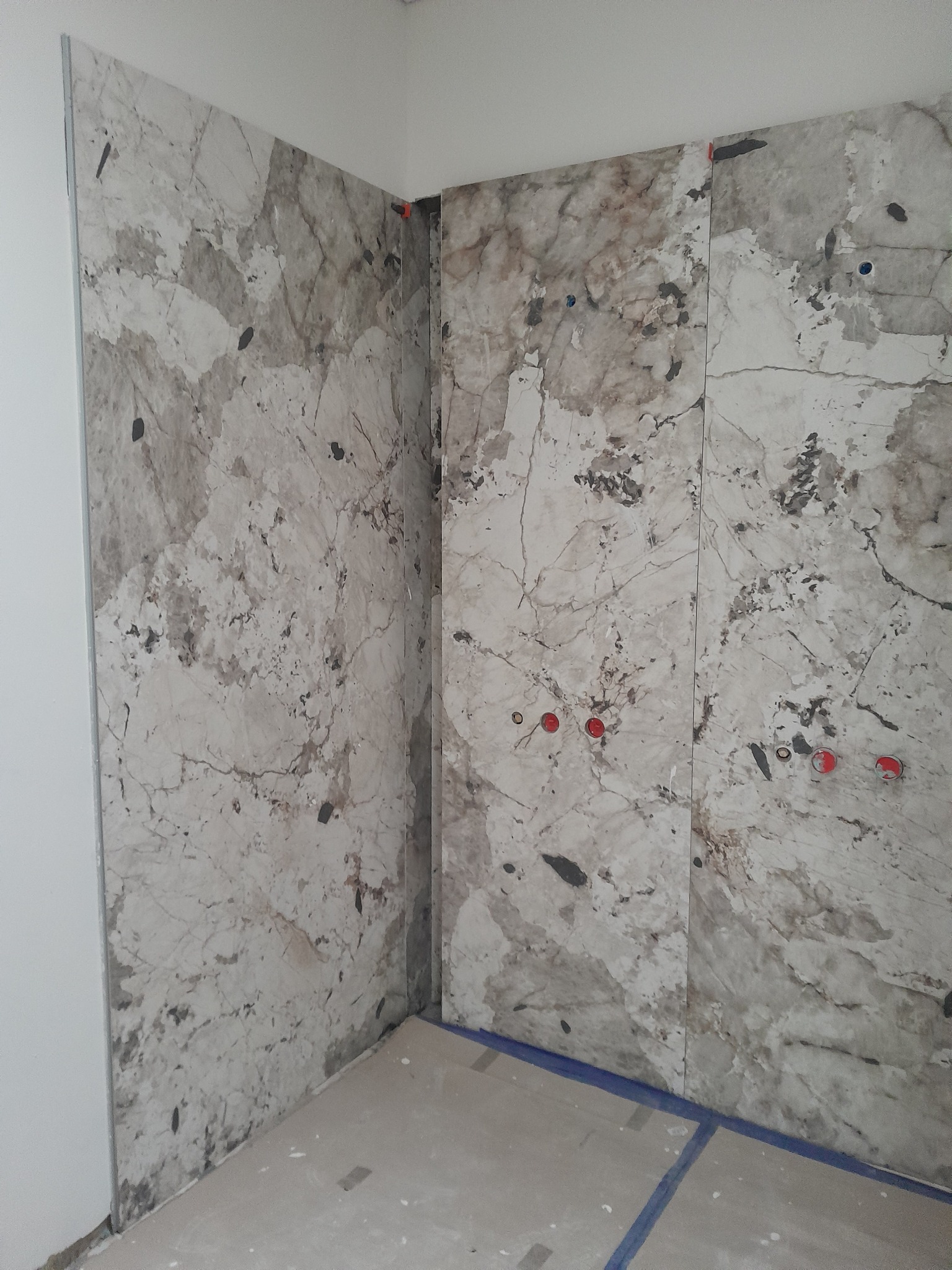 Ściana łazienki wyłożona płytkami imitującymi marmur, z widocznymi otworami na armaturę i niewielkimi elementami poziomowania płytek.