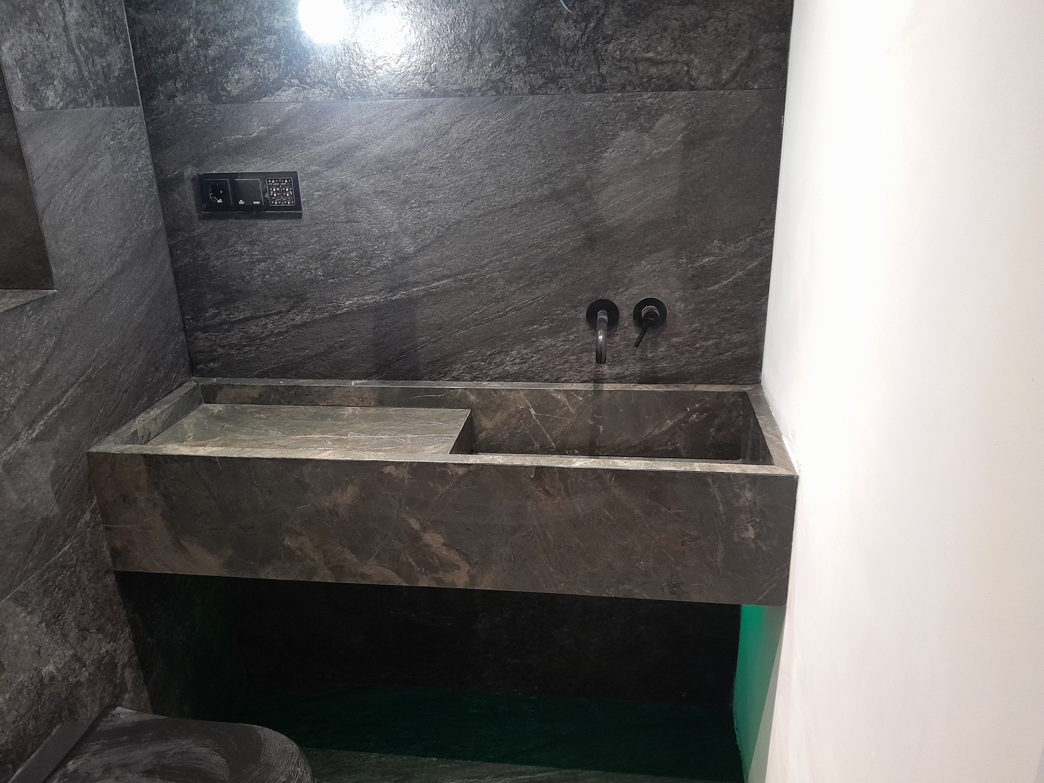 Nowoczesna łazienka z umywalką z ciemnego kamienia z widocznym oświetleniem LED od dołu, dopasowaną armaturą i panelem sterowania na ścianie z kamienia.