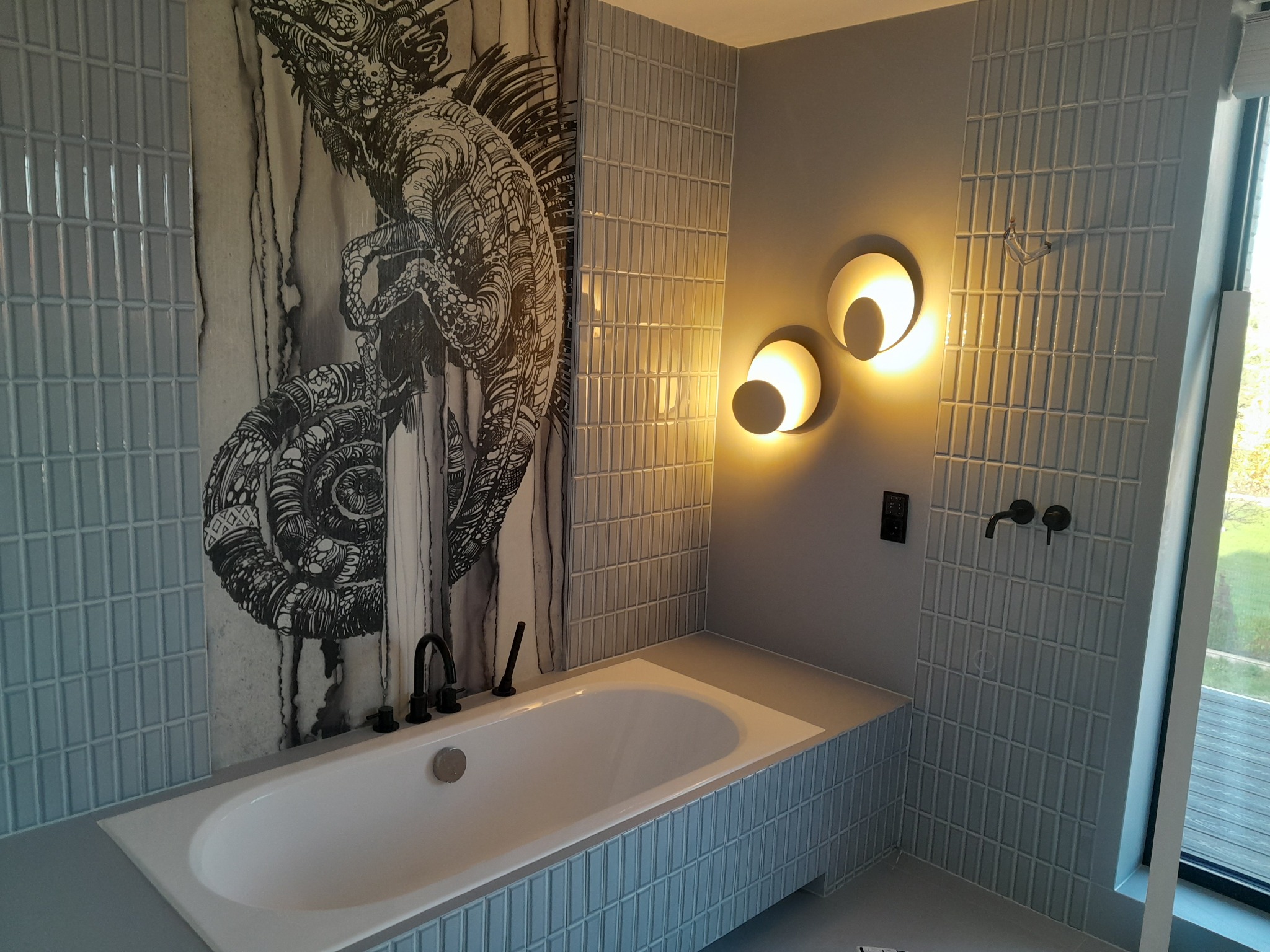 Wykończona łazienka z wanną obłożoną jasnoniebieską, wąską mozaiką, ściana z grafiką kameleona, okrągłe lampy ścienne i czarna armatura.