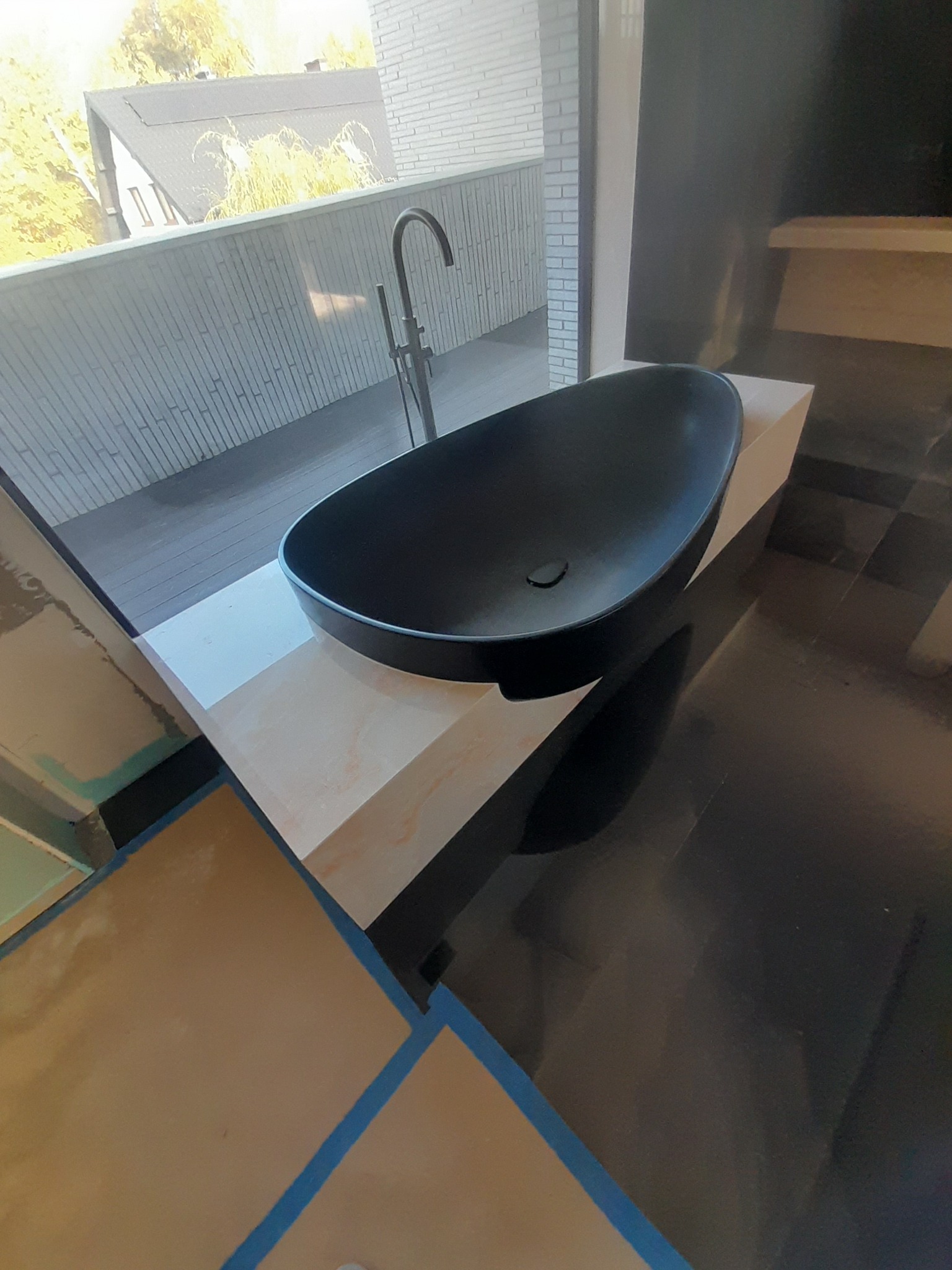 Czarna, owalna umywalka nablatowa z wysoką baterią, ustawiona na blacie z efektem marmuru, widok z góry na tle okna z widokiem na dom i drzewa, podłoga w czarne płyty, pomieszczenie w trakcie prac...