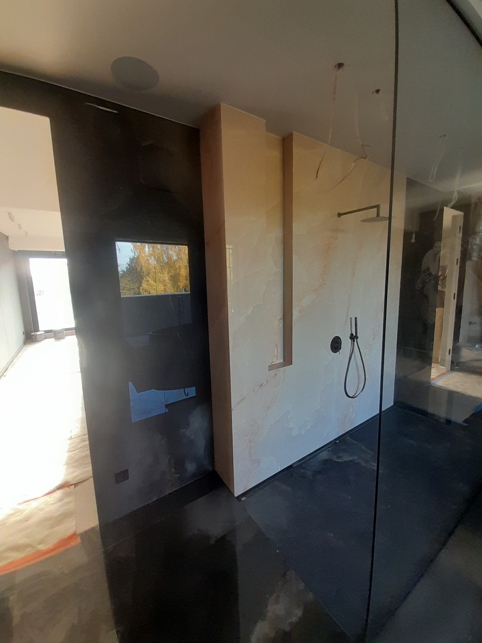 Nowoczesna łazienka w trakcie wykańczania, widoczna kabina prysznicowa ze szklaną ścianą, marmurowe płytki na ścianach i czarna podłoga z połyskiem, częściowo widoczne okno z widokiem na drzewa.