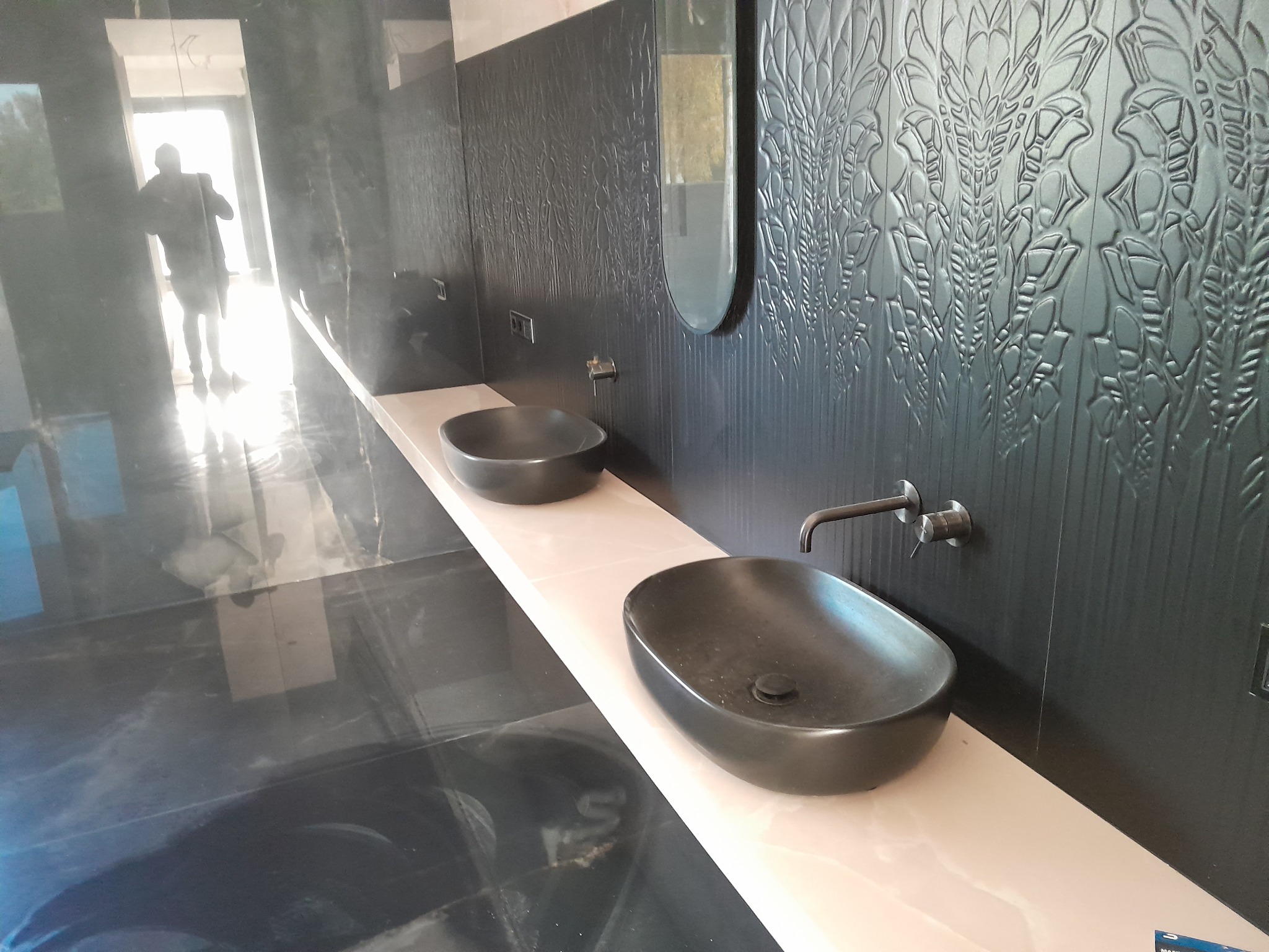 Nowoczesna łazienka w Warszawie z dwoma czarnymi umywalkami na długim, jasnym blacie, ozdobna czarna ściana z motywem roślinnym i podłużnym lustrem, widoczna osoba w tle.