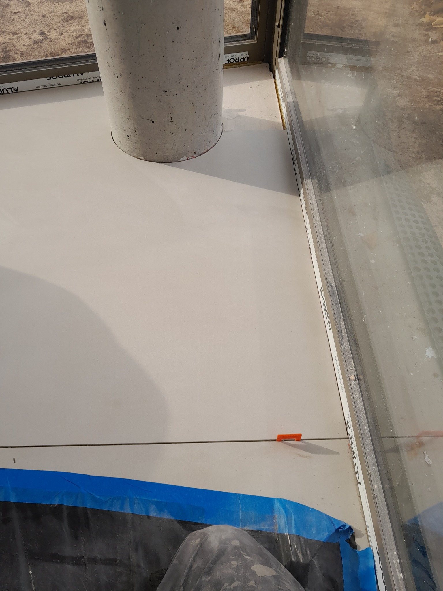 Układanie jasnych płytek podłogowych przy użyciu krzyżyków dystansowych, widoczne zabezpieczenie folią i element architektoniczny w kształcie walca.