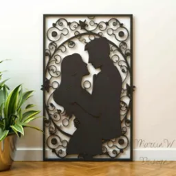 Czarna, ażurowa dekoracja ścienna przedstawiająca sylwetki obejmującej się pary, otoczone roślinnym ornamentem, stojąca na podłodze obok doniczki z rośliną.