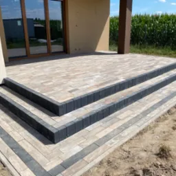 Nowoczesny taras z kostki brukowej w odcieniach beżu i szarości, prowadzący do domu z drewnianą elewacją i dużymi oknami, widok na pole kukurydzy w tle.