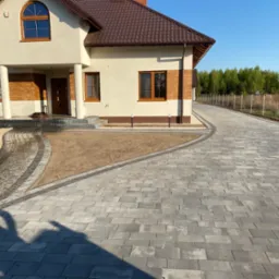 Nowoczesny dom z podjazdem wyłożonym szarą kostką brukową, obrzeża z ciemniejszej kostki, trawnik w trakcie zakładania, oświetlenie wzdłuż ścieżki.