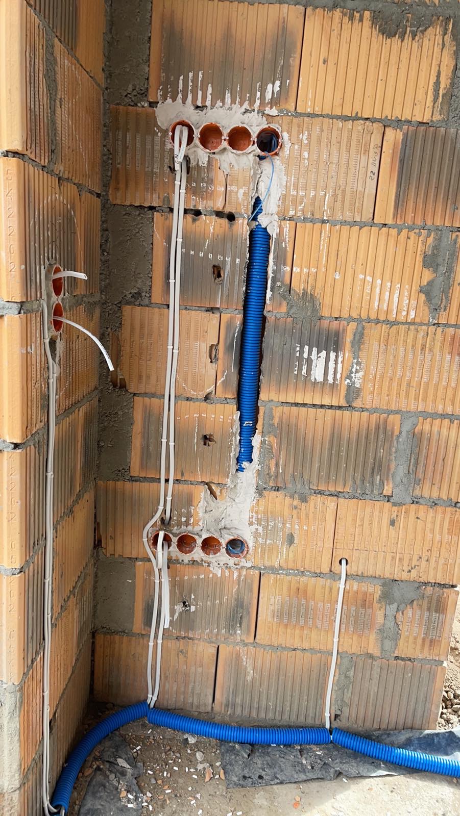 Ściana z cegły z wywierconymi otworami i poprowadzonymi przewodami elektrycznymi w białych rurkach oraz niebieska rura karbowana, widoczne ślady zaprawy.