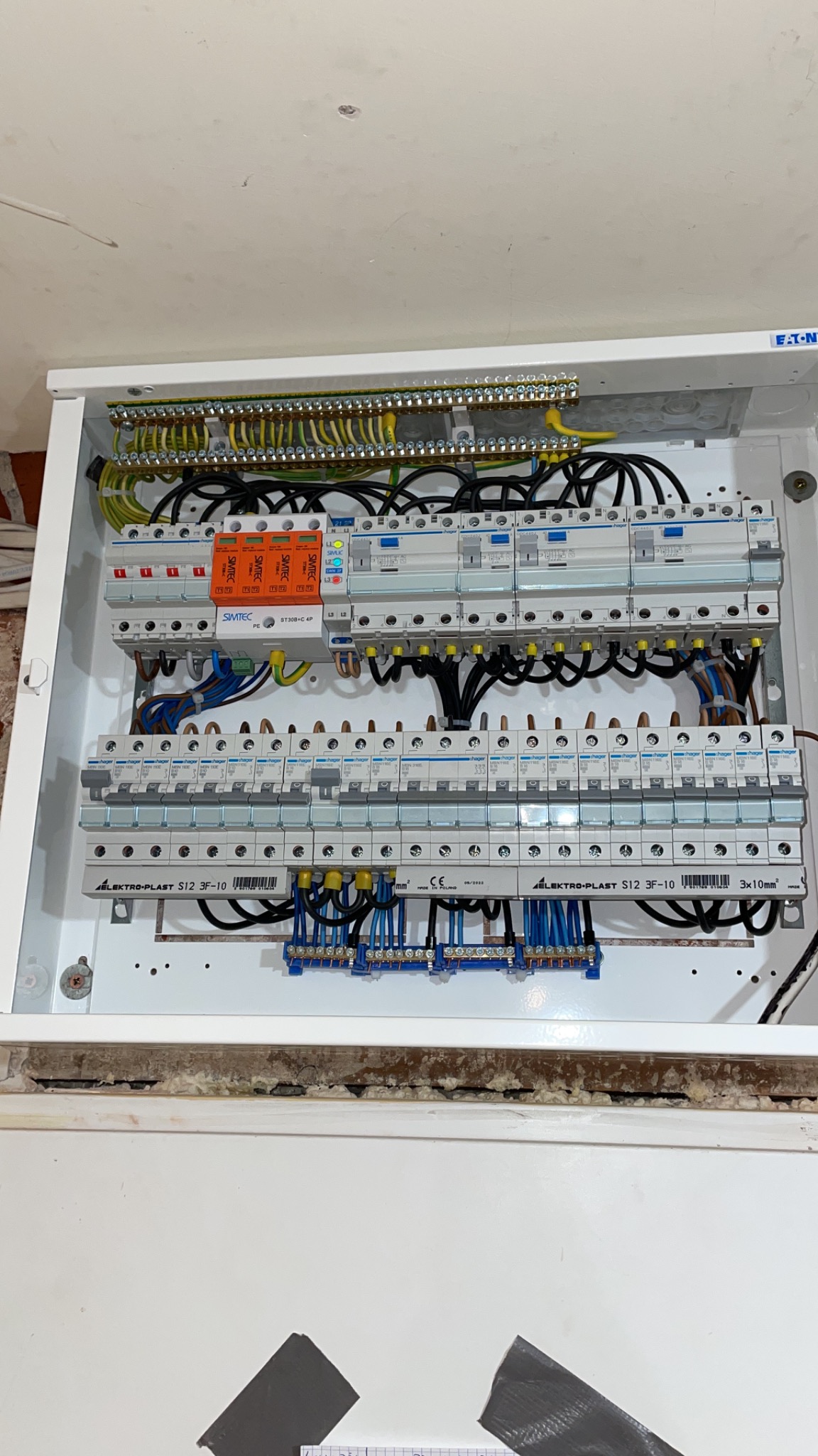 Otwarta, biała skrzynka rozdzielcza z zamontowanymi bezpiecznikami, listwami zaciskowymi i przewodami. Widoczne marki Simtec i Elektro-Plast. Częściowo uszczelniona obudowa z widocznymi śladami...