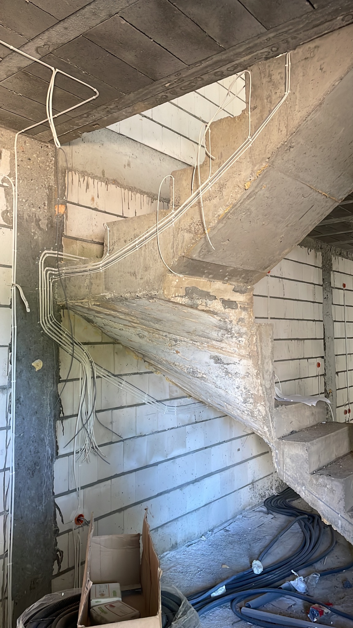 Instalacja elektryczna w surowym wnętrzu: białe przewody prowadzone wzdłuż betonowych schodów i ściany z bloczków, widoczne kable w peszlu na podłodze i kartonowe pudło z elementami instalacji.