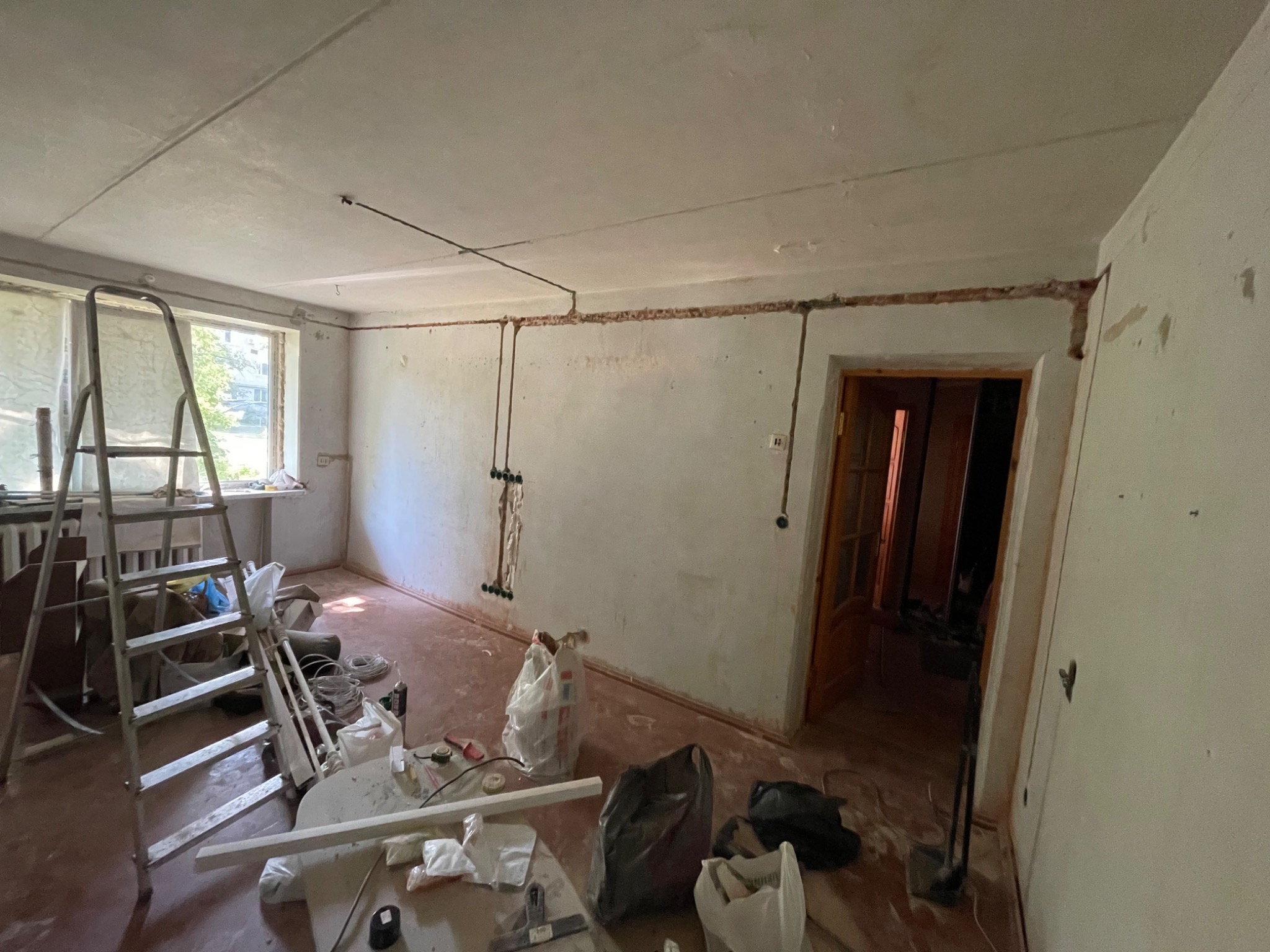 Pokój w trakcie remontu, z widocznymi bruzdami w ścianach pod instalację elektryczną, drabina oparta o ścianę przy oknie, narzędzia i materiały budowlane na podłodze.
