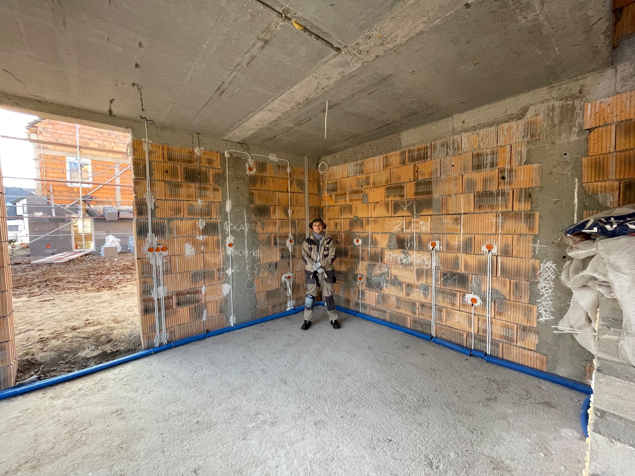 Elektryk w kombinezonie stoi w surowym pomieszczeniu z ceglanymi ścianami, na których widoczne są instalacje elektryczne w rurkach i puszki pod gniazdka, z niebieską rurą ochronną przy podłodze,...