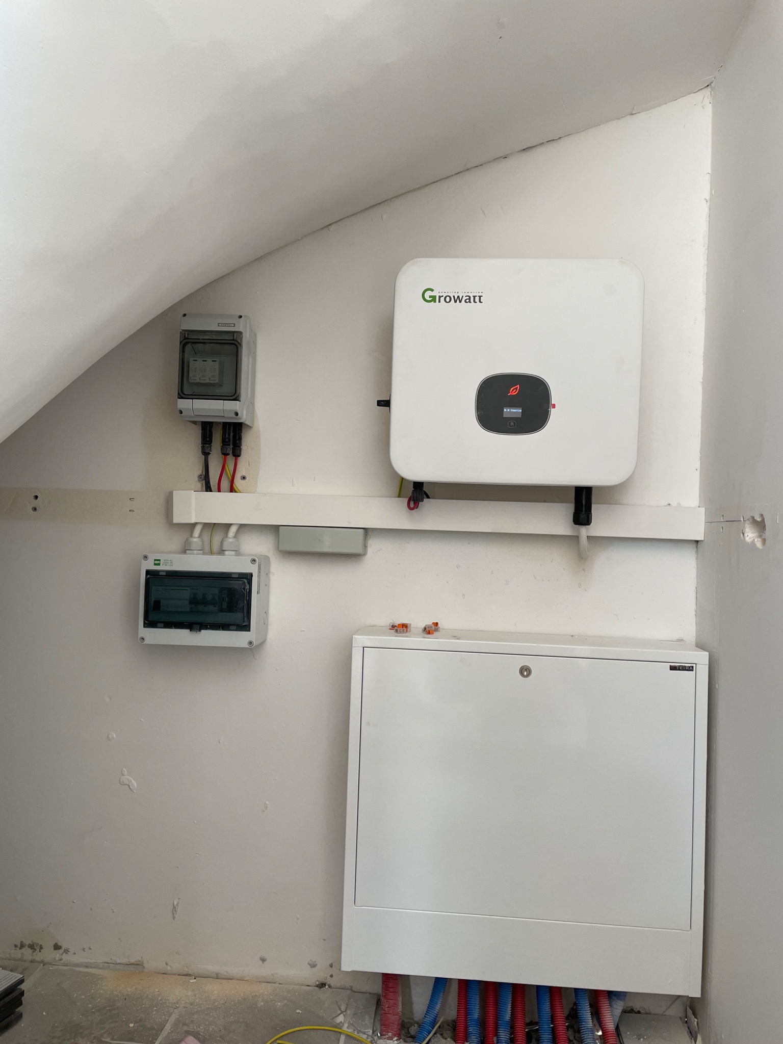 Biała skrzynka elektryczna z podłączonymi niebieskimi i czerwonymi rurami, obok zamontowany inwerter marki Growatt, dwa liczniki elektryczne na ścianie w surowym stanie.
