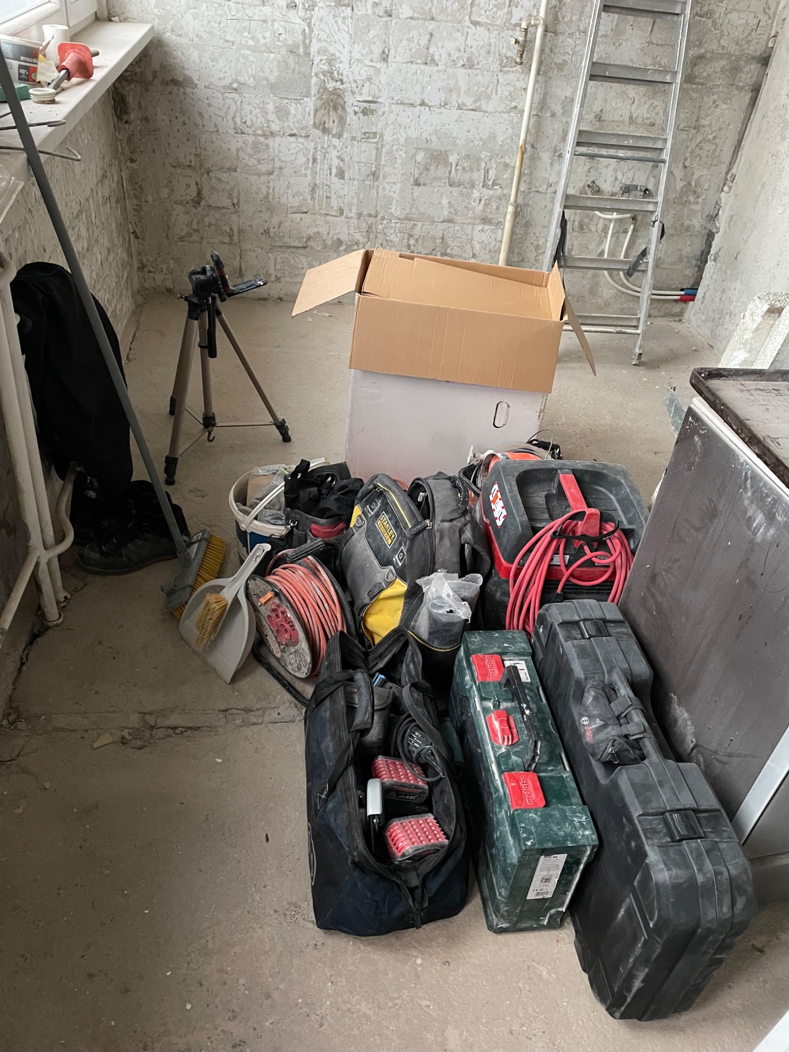 Stos narzędzi i sprzętu elektrycznego, w tym torby narzędziowe, odkurzacz przemysłowy i drabina, w surowym pomieszczeniu z nieotynkowanymi ścianami.