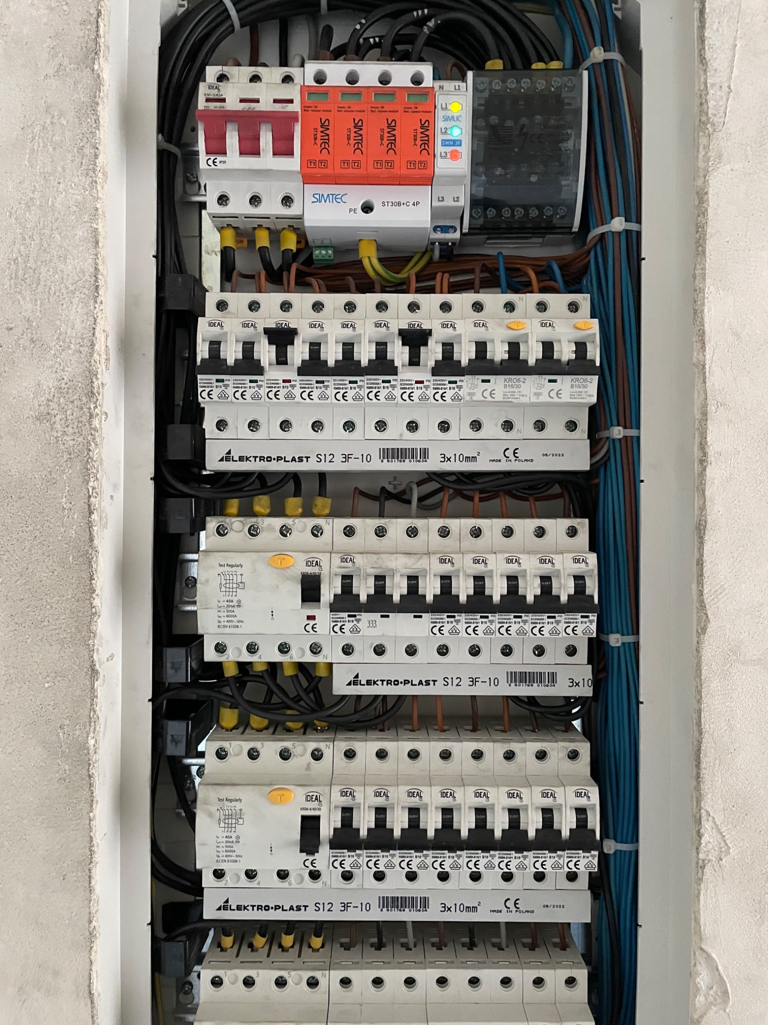Rozdzielnica elektryczna z zainstalowanymi bezpiecznikami, wyłącznikami różnicowoprądowymi i ogranicznikami przepięć marki Simtec, widoczne przewody i oznaczenia.