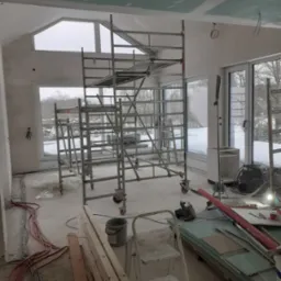 Wnętrze w trakcie prac wykończeniowych: rusztowanie, płyty gipsowo-kartonowe, kable elektryczne i widok zimowego krajobrazu za oknem.
