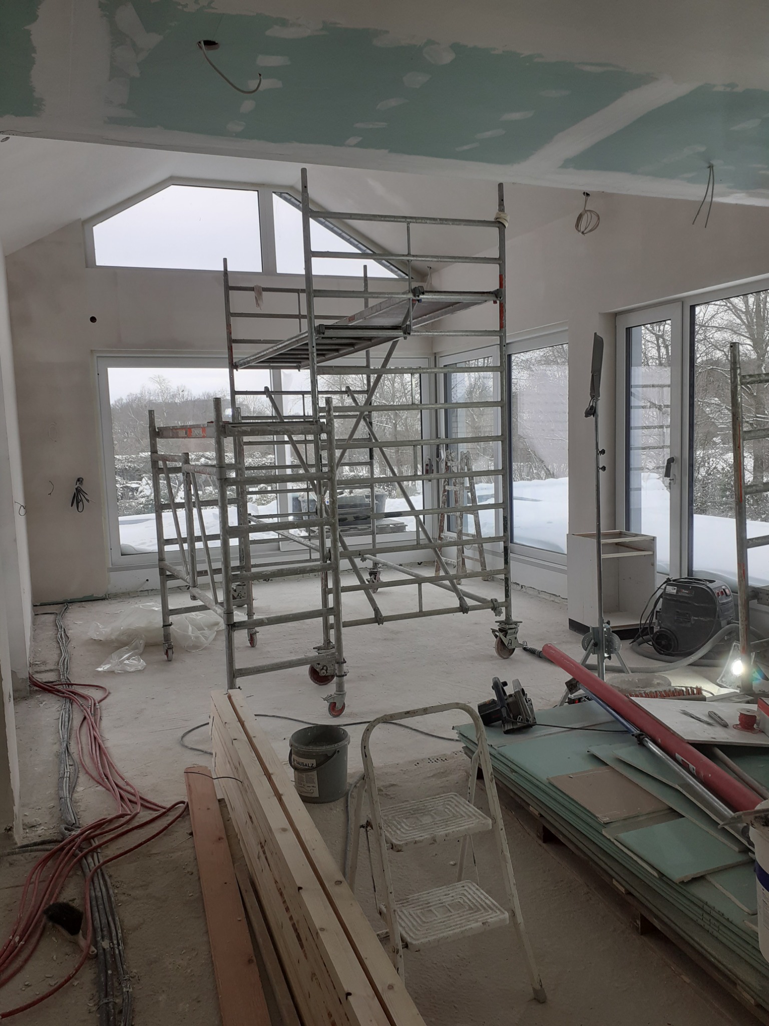 Wnętrze w trakcie prac wykończeniowych: rusztowanie, płyty gipsowo-kartonowe, kable elektryczne i widok zimowego krajobrazu za oknem.