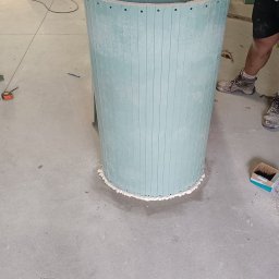 Perfect Wall - Wznoszenie okrągłej kolumny z płyt kartonowo-gipsowych, zabezpieczonej pianką montażową u podstawy, widoczne śruby mocujące i buty robocze wykonawcy w tle.