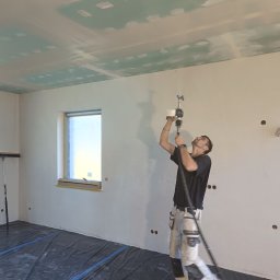 Perfect Wall - Pracownik w trakcie nakładania gładzi natryskowej na sufit z płyt kartonowo-gipsowych w pomieszczeniu z zabezpieczoną folią podłogą i oknem.