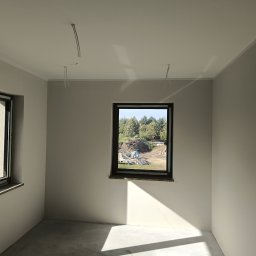 Perfect Wall - Wnętrze pokoju po remoncie, świeżo pomalowane ściany w odcieniach szarości, widoczne dwa okna z widokiem na zewnątrz, na suficie wystające przewody elektryczne, na zewnątrz budynku widoczny plac...