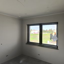 Perfect Wall - Wnętrze pokoju po szpachlowaniu i malowaniu ścian na jasny odcień, widoczne okna z parapetami, wystające kable oświetleniowe i zaklejone okno folią ochronną.