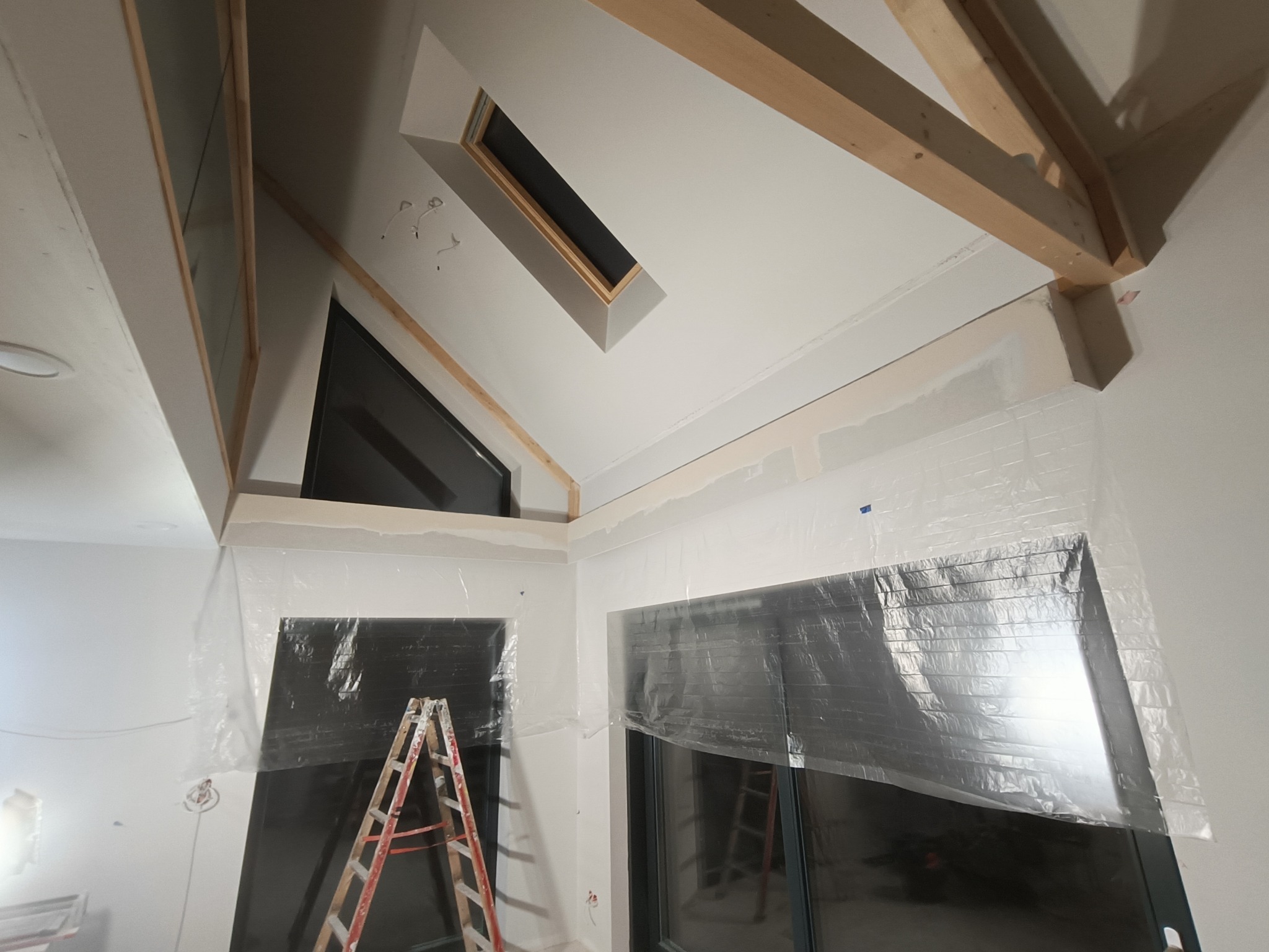 Wnętrze w trakcie prac wykończeniowych: zabezpieczone folią okna, widoczna drabina, surowy stan ścian i sufitu z elementami drewnianymi, okno dachowe.