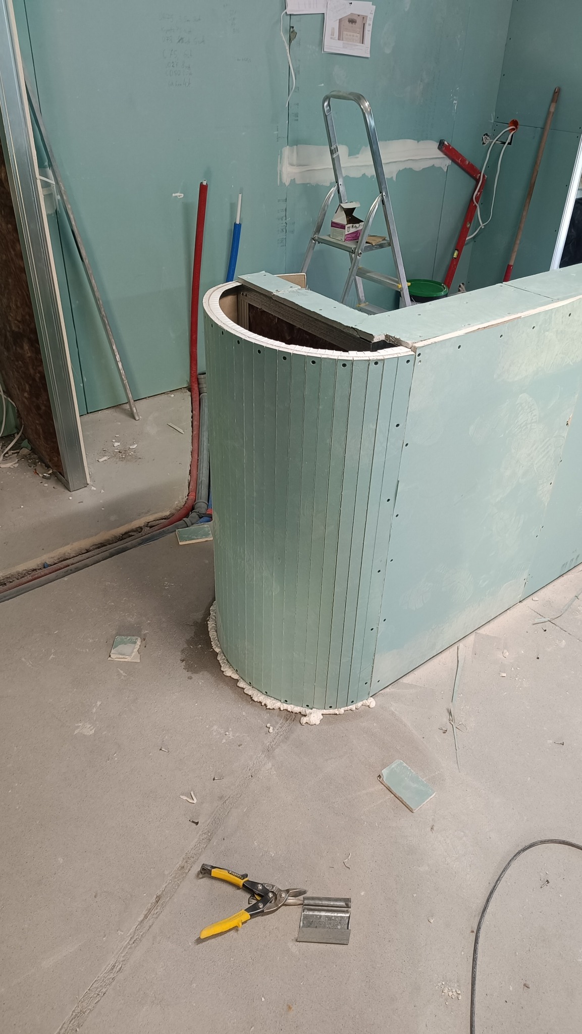 Konstrukcja baru z płyty karton gips, widoczne profile i narzędzia na surowym betonie, w tle ściana z zapiskami i drabina