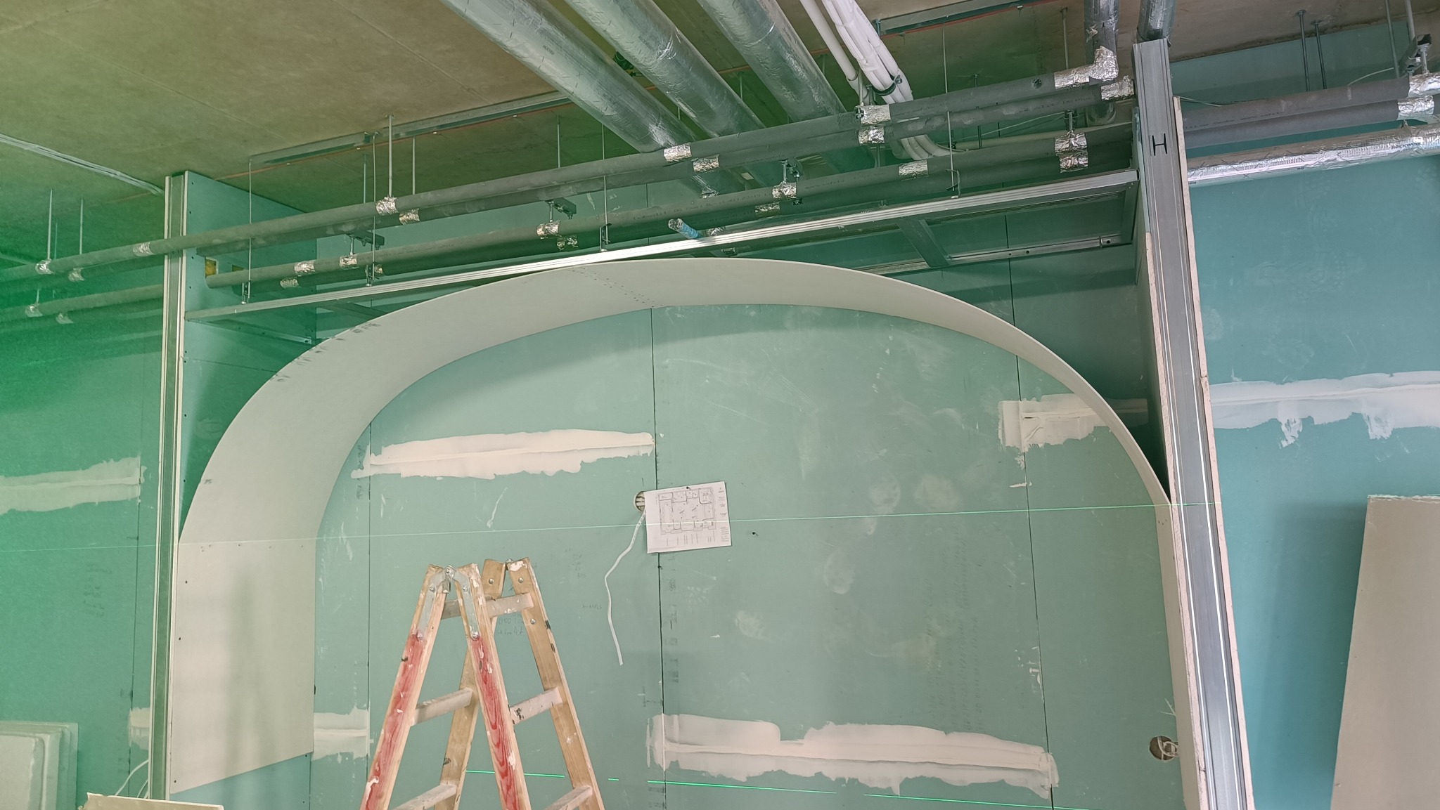 Montaż łuku z płyty kartonowo-gipsowej na ścianie z widocznymi profilami, rurami pod sufitem i laserowym poziomowaniem. Na ścianie przyklejony schemat.