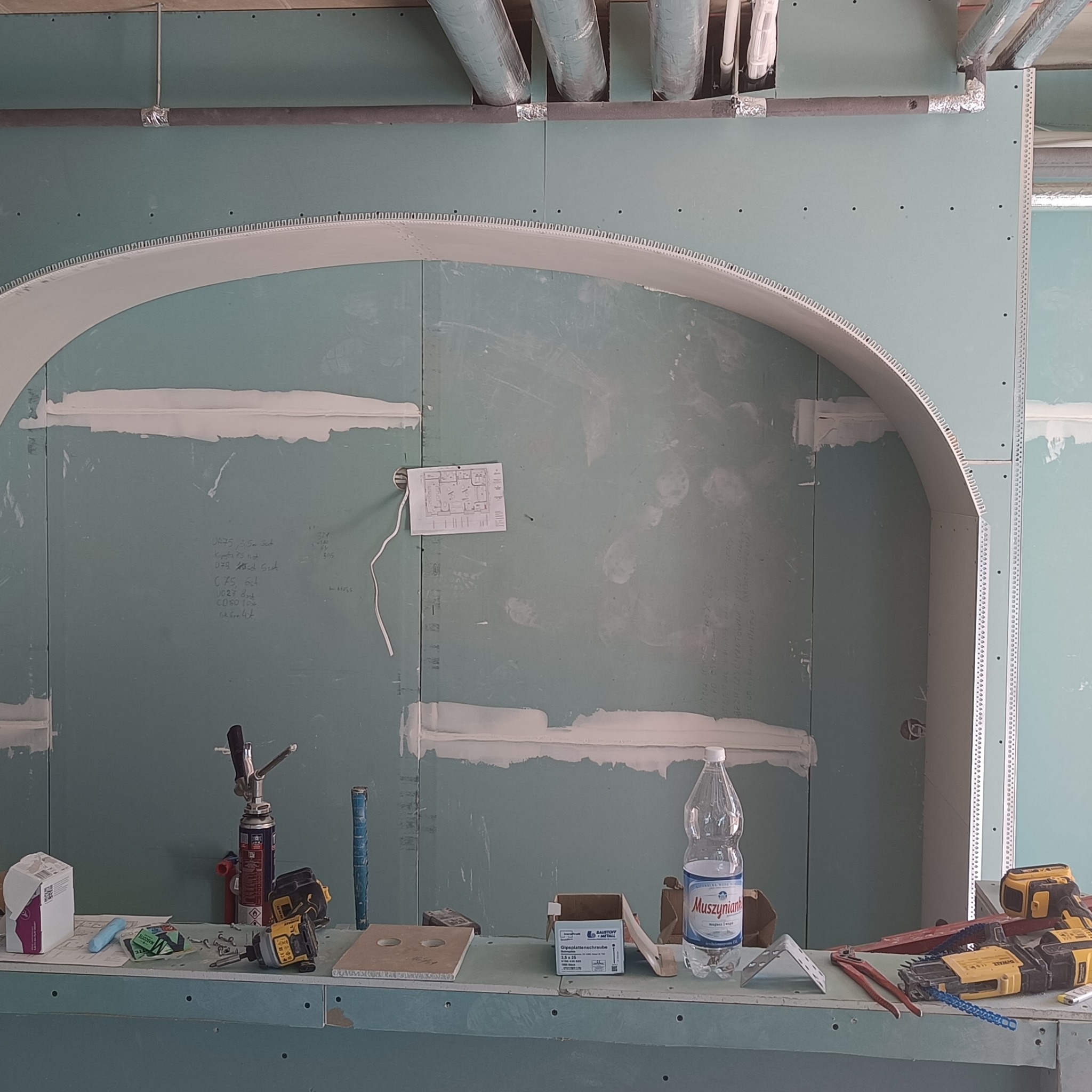 Montaż płyty kartonowo-gipsowej z wykończeniem łukowym, widoczne narzędzia i plan techniczny na ścianie.
