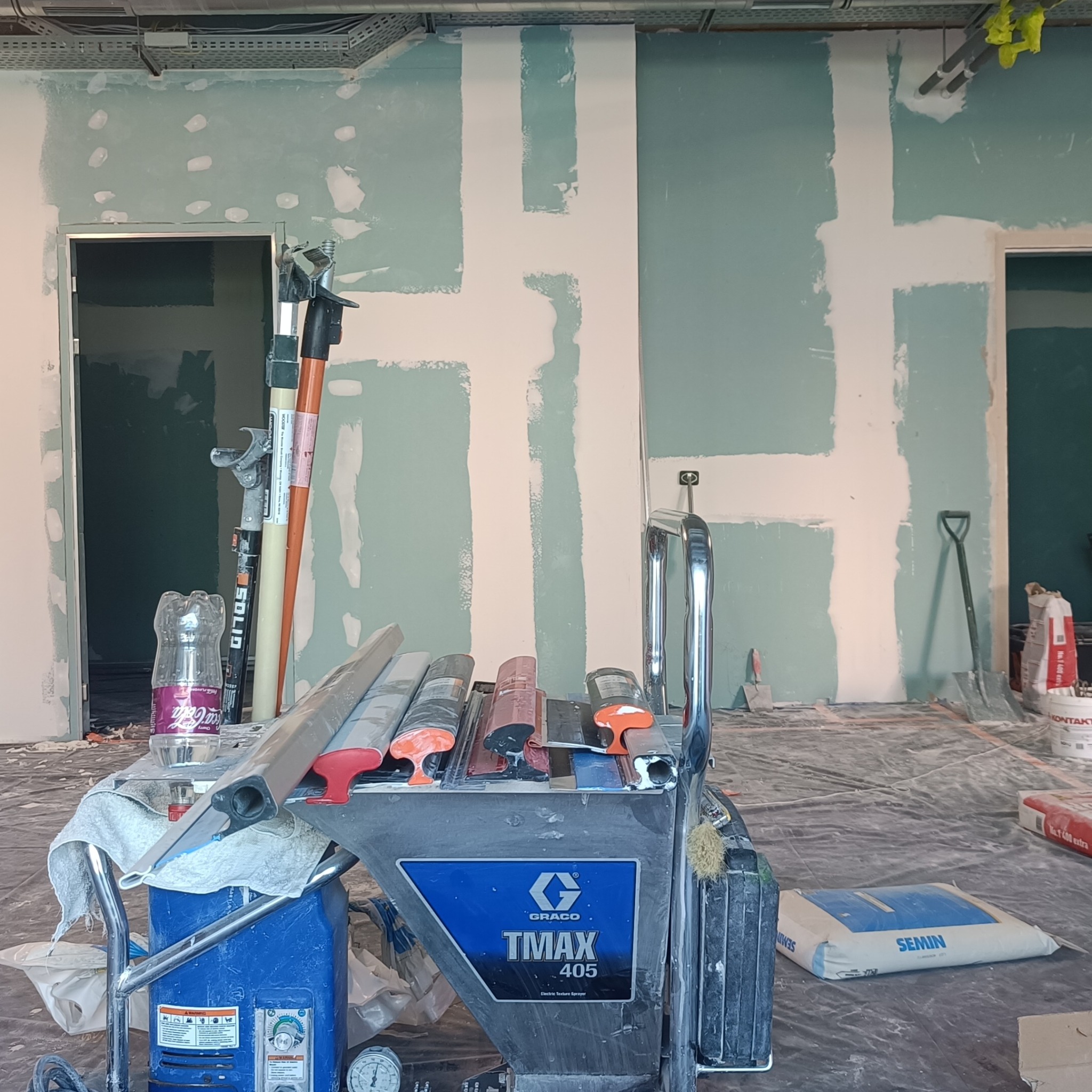 Wnętrze w trakcie prac przygotowawczych we Wrocławiu: ściany po wstępnym szpachlowaniu, widoczny agregat szpachlarski Graco TMAX 405, narzędzia, worki z gładzią i butelka Coca-Coli.