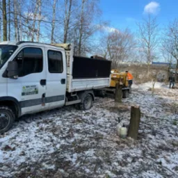 Biały samochód dostawczy z logiem firmy ogrodniczej na tle zimowego krajobrazu z pracownikami przy rębaku i wycince drzew