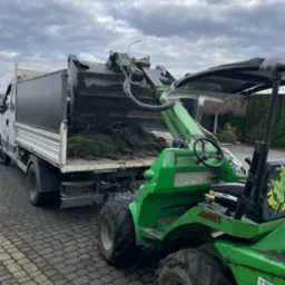 Zielona ładowarka Avant 635 z łyżką załadowuje darń do skrzyni ładunkowej białej ciężarówki na brukowanym podjeździe, pochmurne niebo w tle.