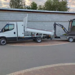 Biały samochód dostawczy z wywrotką i przyczepą transportuje żółto-szarą mini koparkę Volvo EC18E na brukowanym placu, na tle szarego muru z betonowych bloków.