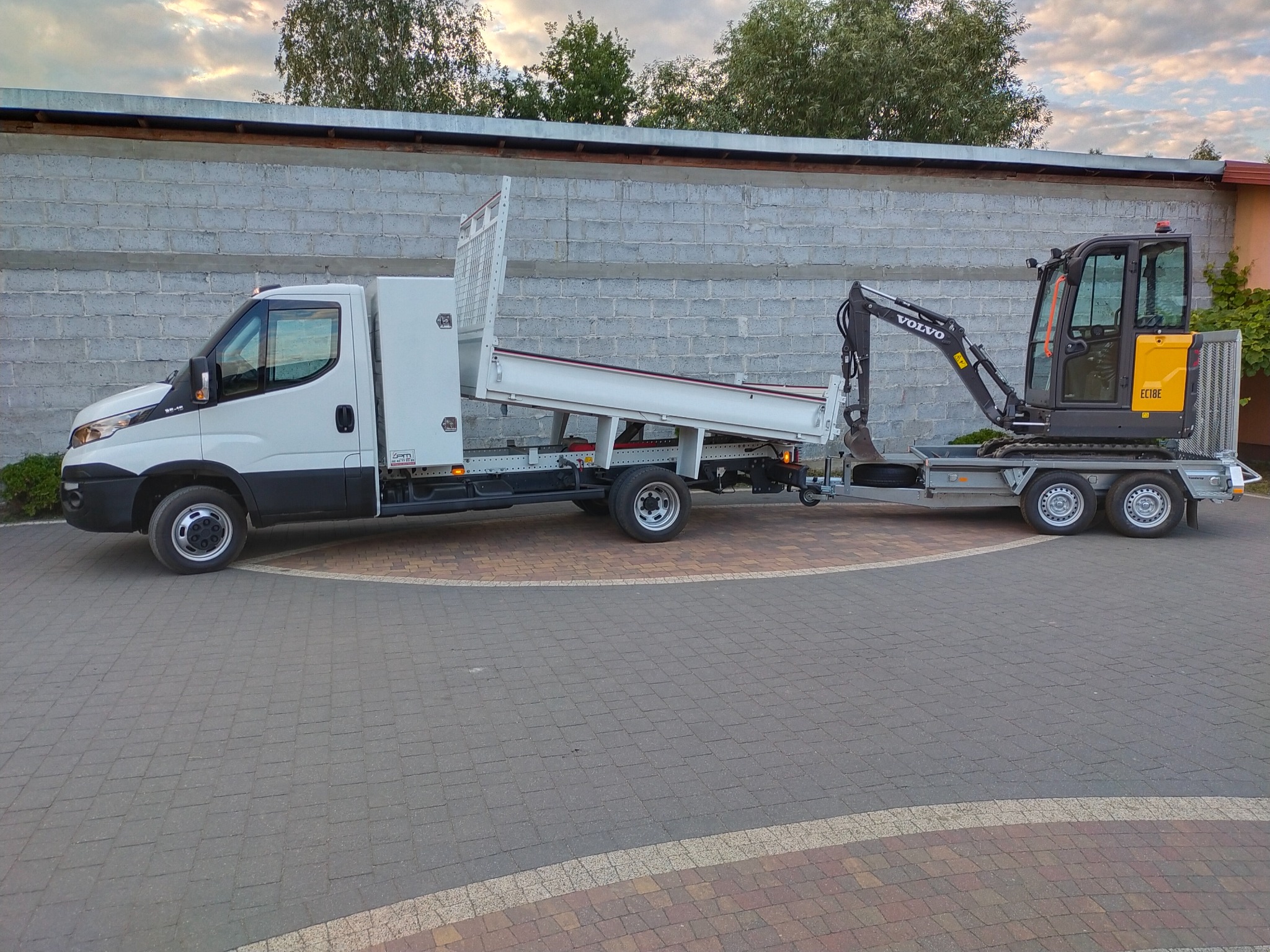 Biały samochód dostawczy z wywrotką i przyczepą transportuje żółto-szarą mini koparkę Volvo EC18E na brukowanym placu, na tle szarego muru z betonowych bloków.