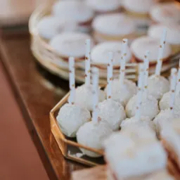 Białe cake pops z posypką w złotej, geometrycznej tacy, obok ciasteczka na złotym paterze, na drewnianym stole.