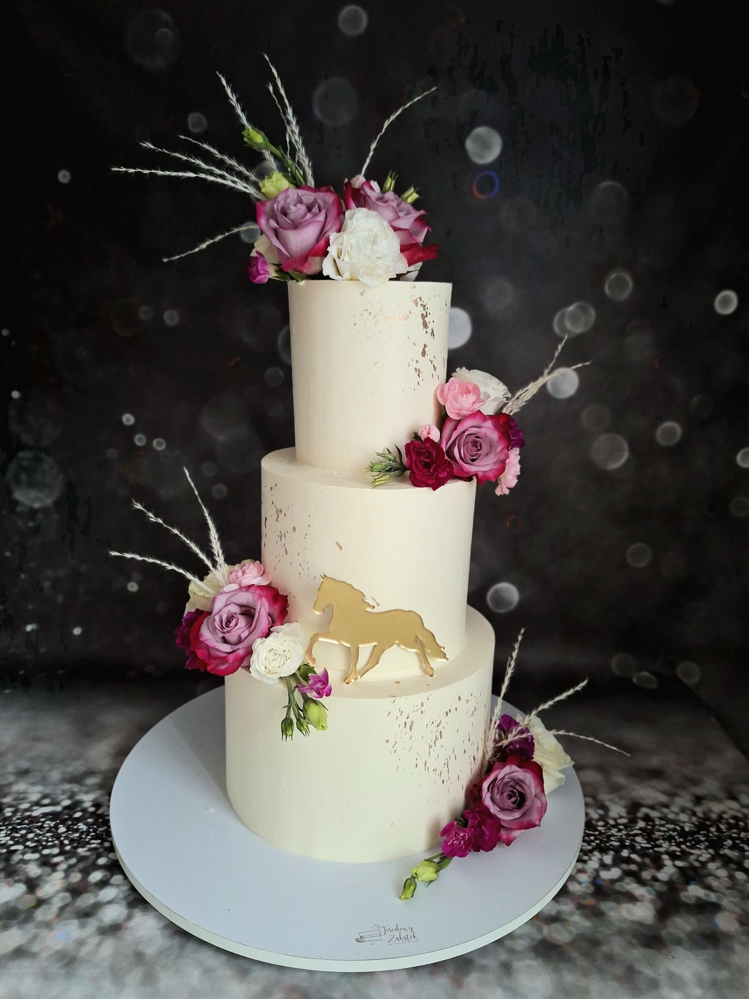 Trzypiętrowy tort weselny w kremowym kolorze, ozdobiony złotymi plamkami i kompozycjami z różowych, białych i bordowych róż oraz złotą figurką konia.