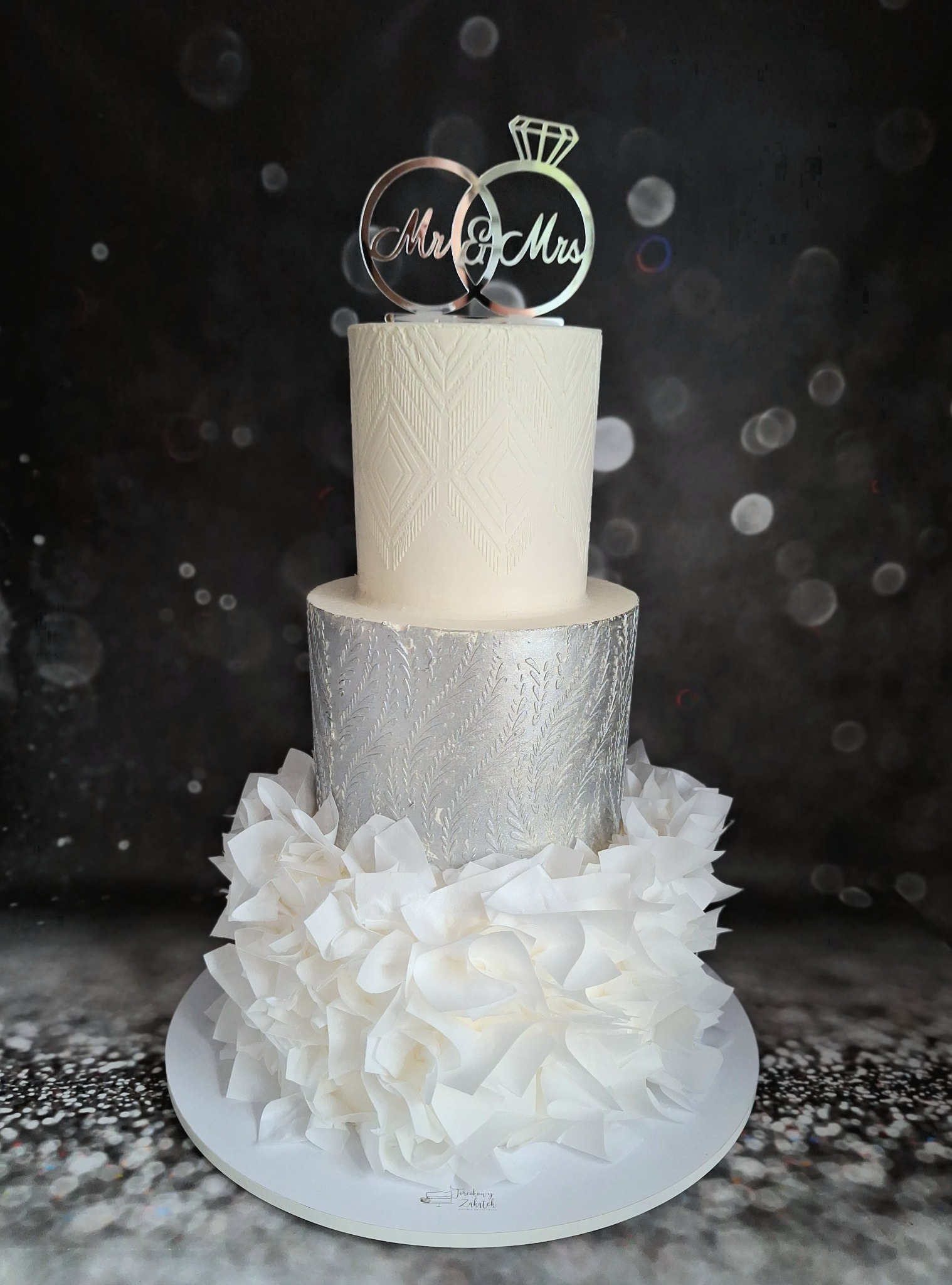 Trzypiętrowy tort weselny w odcieniach bieli i srebra, zdobiony wzorami geometrycznymi i falbanami z papieru, z topperem 'Mr & Mrs' z pierścionkami na czarnym tle z bokeh.