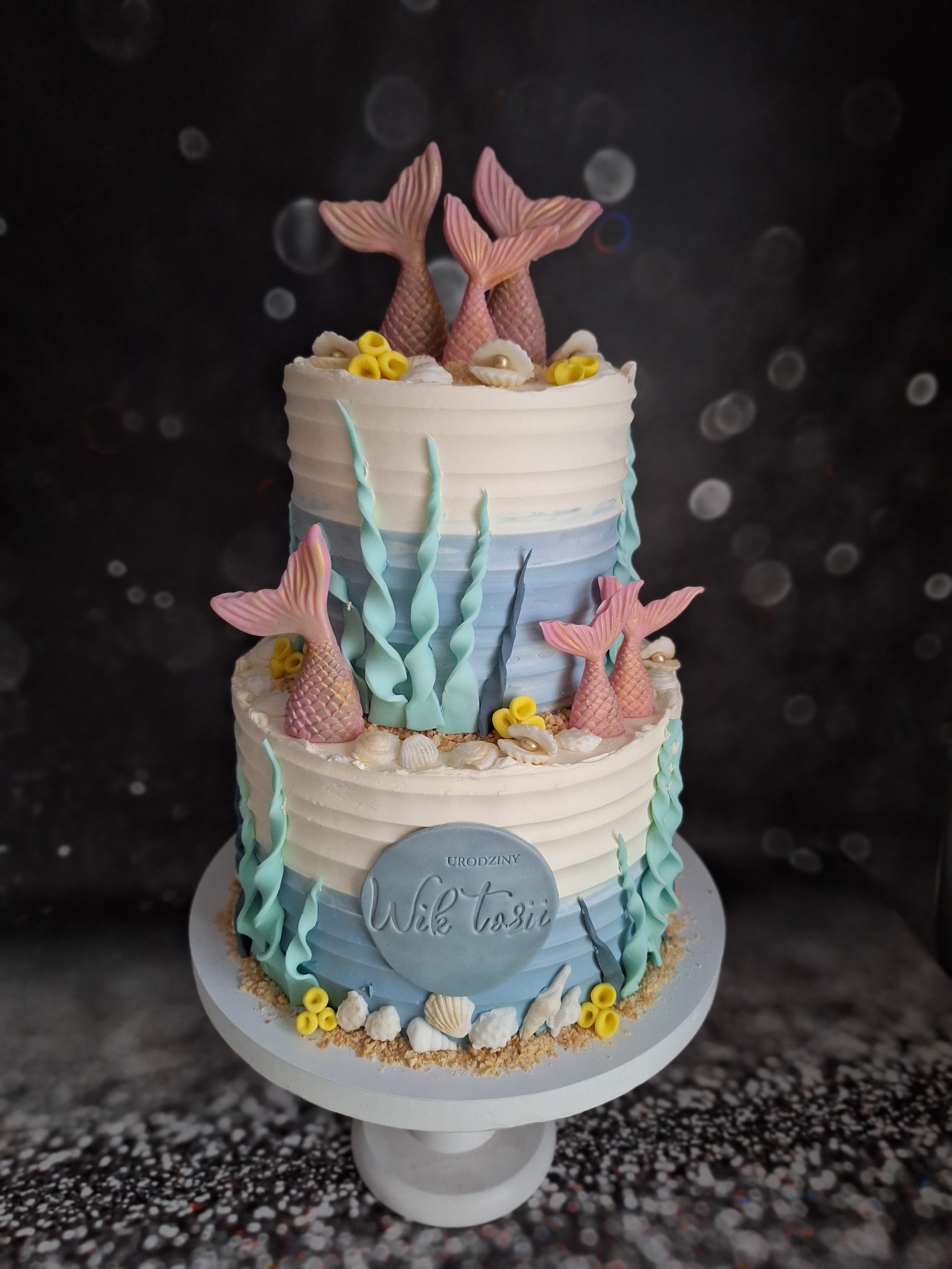 Dwupiętrowy tort urodzinowy w stylu morskim, zdobiony kremem, falistymi algami, muszelkami i różowymi ogonami syren, umieszczony na białym stojaku na tle ciemnego, rozmytego tła.