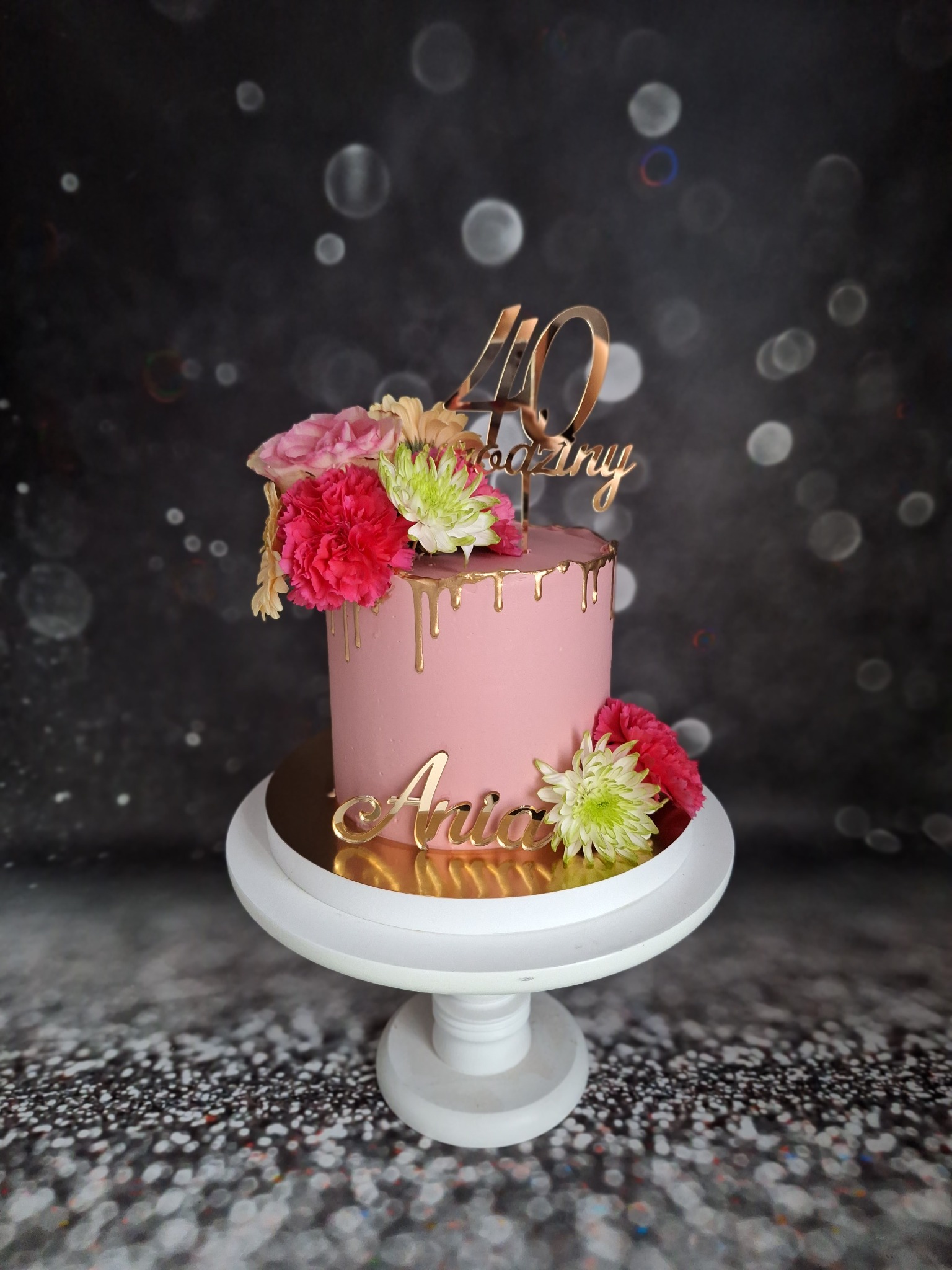Różowy tort urodzinowy z kwiatami i złotymi akcentami, dekorowany napisem '40 Amazing' i imieniem 'Ania', umieszczony na białym stojaku na tle czarnego tła z efektem bokeh.