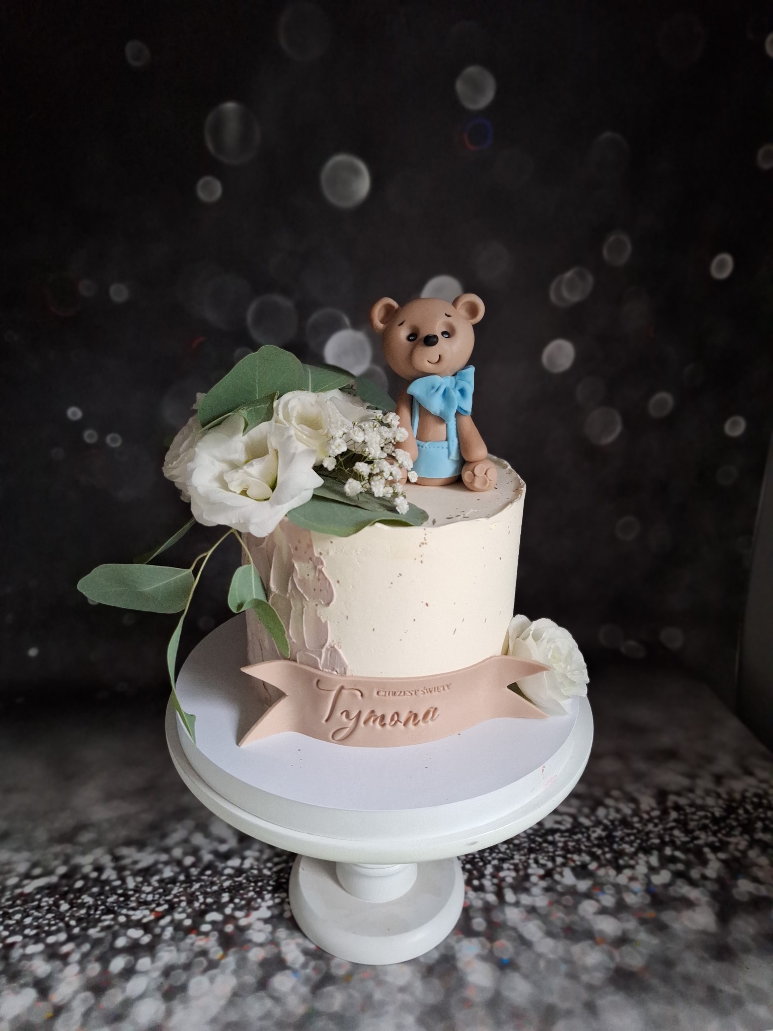 Tort okolicznościowy z kremowym lukrem, kwiatami i figurką misia w błękitnej muszce, umieszczony na białym stojaku na tle ciemnego, błyszczącego tła.