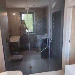 Pawel Grzywa - Nowoczesna łazienka z kabiną prysznicową typu walk-in z ciemnego szkła, mozaiką na ścianach, wiszącą miską WC i umywalką nablatową, widok przez szklaną ścianę na zieleń za oknem.