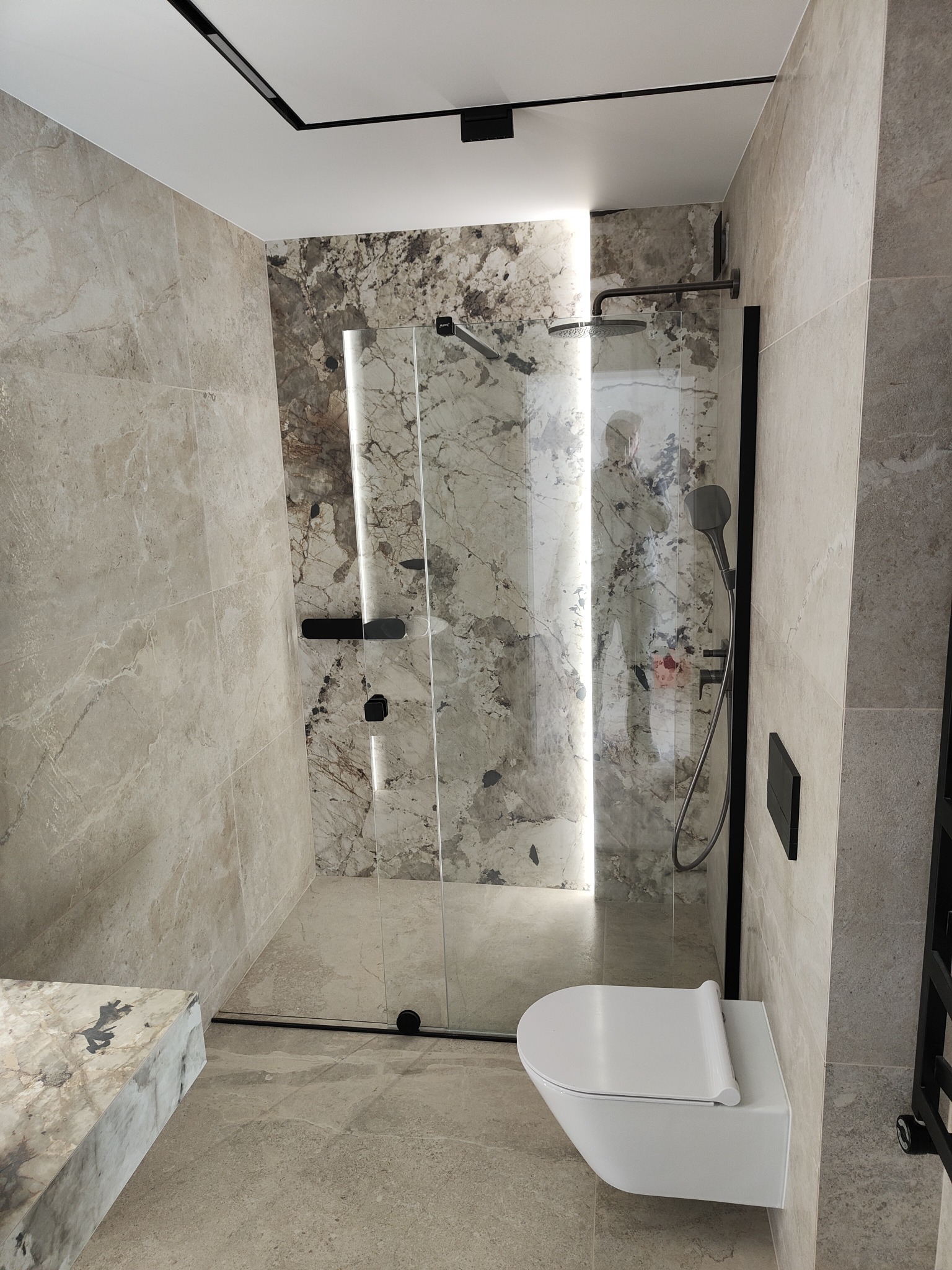 Nowoczesna łazienka z kabiną prysznicową typu walk-in, wykończona płytkami imitującymi marmur w odcieniach beżu i szarości, z czarnymi akcentami w postaci ramy kabiny i oświetlenia szynowego.