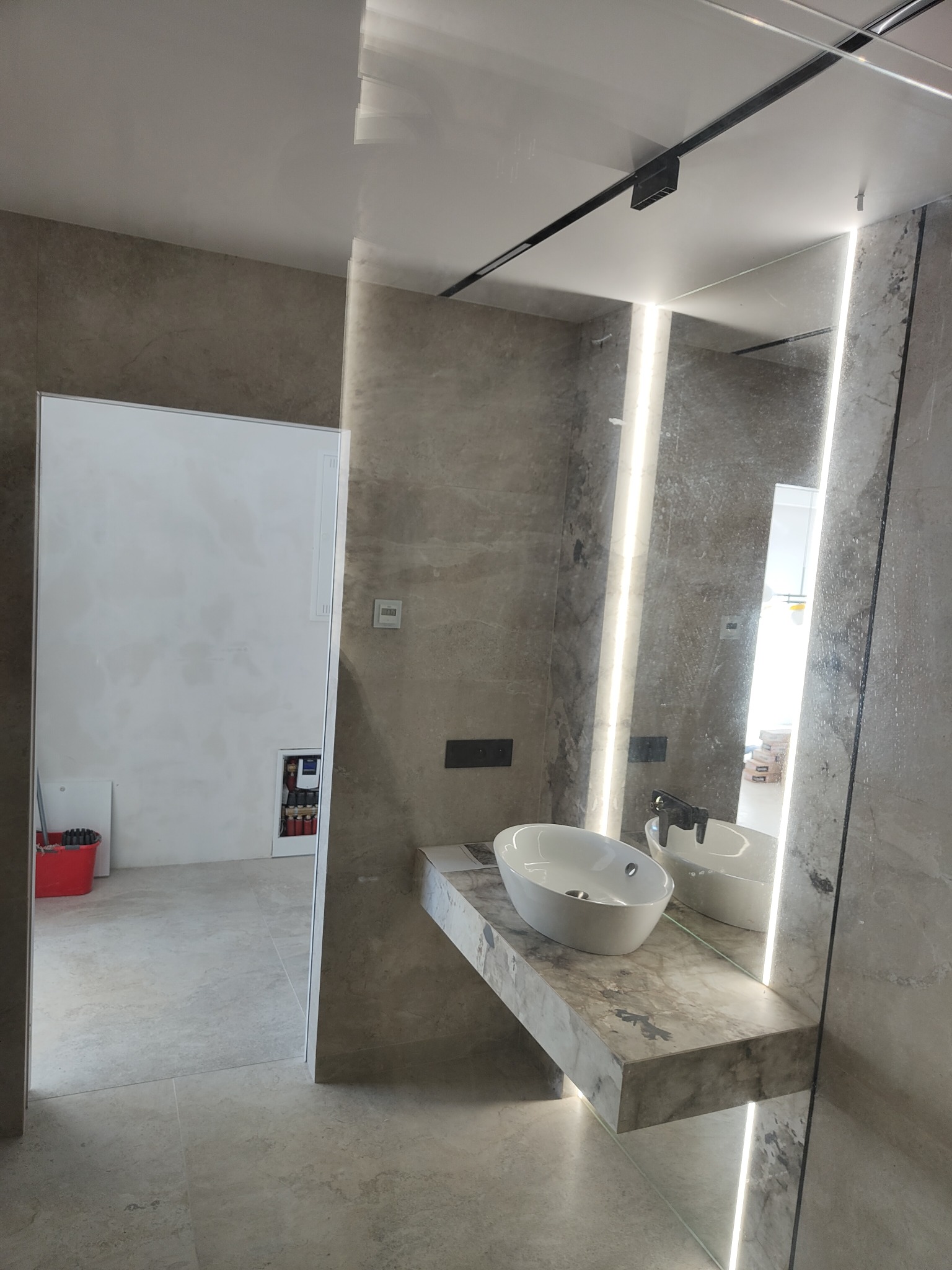 Nowoczesna łazienka z umywalką nablatową na marmurowym blacie, podświetlane lustro, płytki imitujące kamień, widoczna instalacja w szafce i wiadro z mopem w tle.