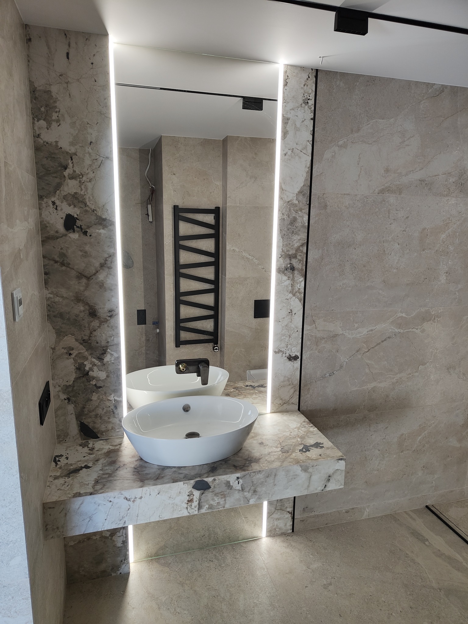 Nowoczesna łazienka z dwoma białymi umywalkami nablatowymi na marmurowym blacie, podświetlane lustro z oświetleniem LED, grzejnik drabinkowy w tle, płytki imitujące marmur na ścianach i podłodze...