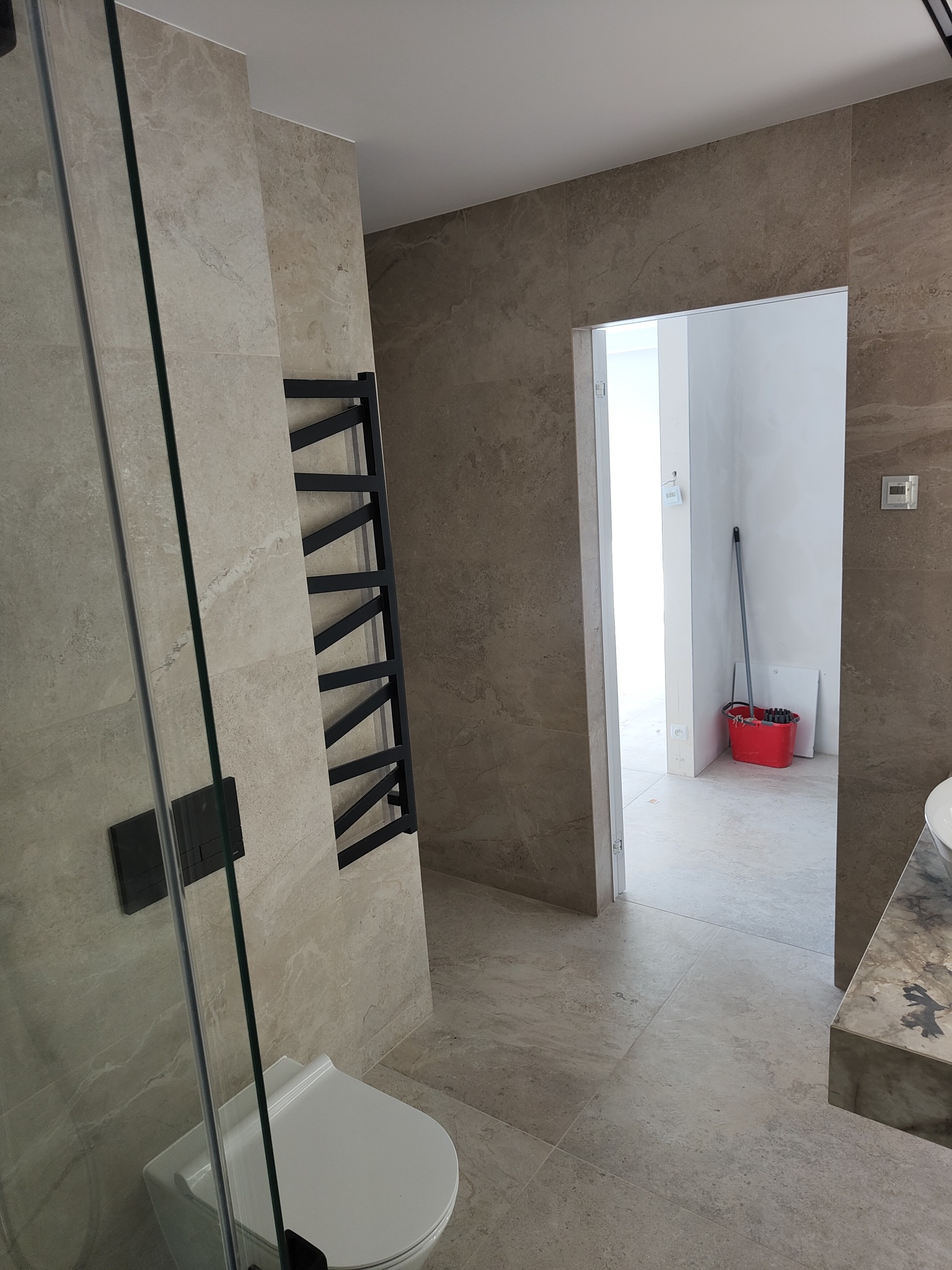 Beżowa łazienka z płytkami imitującymi kamień, czarnym grzejnikiem drabinkowym i kabiną prysznicową ze szkła, widok na sąsiednie pomieszczenie z czerwonym wiadrem i mopem.