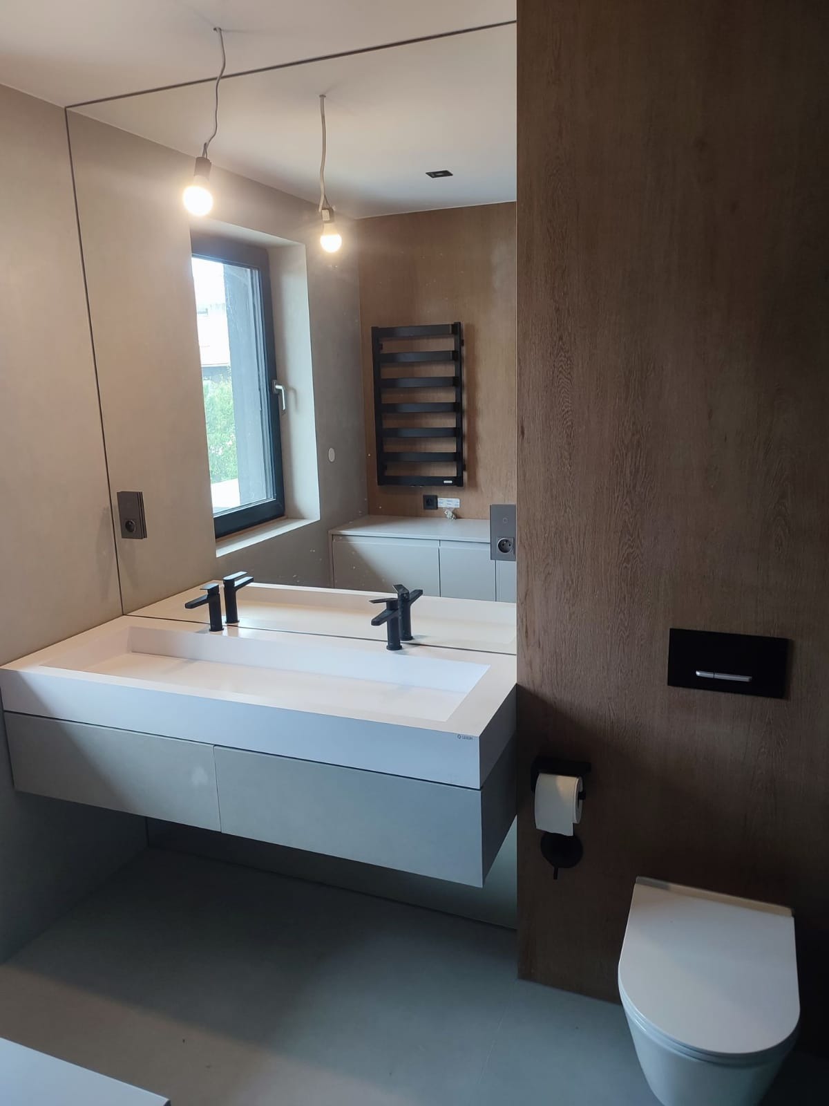 Nowoczesna łazienka z podwójną umywalką nablatową, czarną armaturą i grzejnikiem drabinkowym, oświetlona wiszącymi żarówkami bez kloszy.