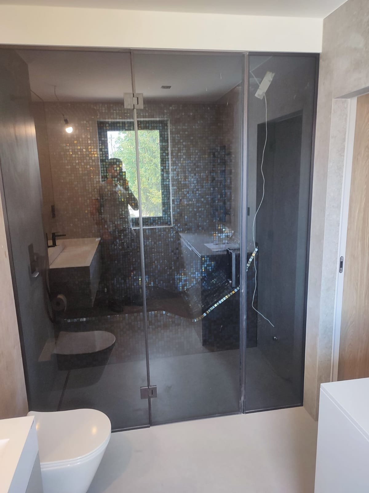 Nowoczesna łazienka z kabiną prysznicową typu walk-in z ciemnego szkła, mozaiką na ścianach, wiszącą miską WC i umywalką nablatową, widok przez szklaną ścianę na zieleń za oknem.