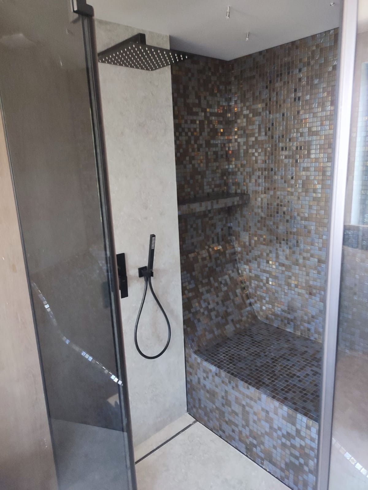 Nowoczesna kabina prysznicowa z deszczownicą i siedziskiem wyłożona mozaiką w odcieniach brązu i szarości, widok z boku przez szklane drzwi.